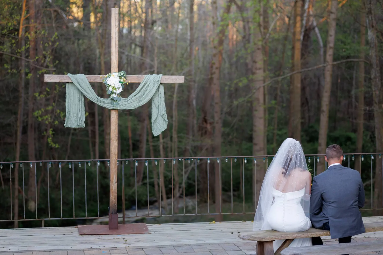 Bride and groom sitting together on deck bench beside the ceremony cross at The Shed at Papa's Creek, Lavonia Georgia